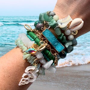 May include: A stack of bracelets featuring green, blue, and white stones, shells, and gold accents. The bracelets are layered on a person's wrist.