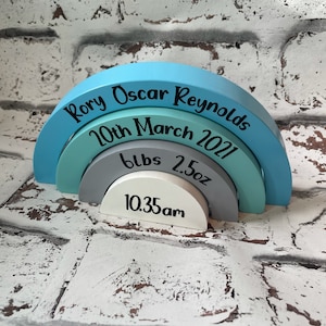 May include: A set of four wooden rainbow arches in shades of blue and grey. The top arch is blue and reads "Rory Oscar Reynolds 10th March 2021". The second arch is teal and reads "6lbs 2.5oz". The third arch is grey and reads "10.35am". The bottom arch is white.