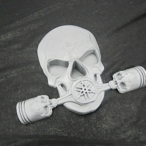 May include: A silver skull with a Yamaha logo on the front and two piston-shaped skulls on either side.