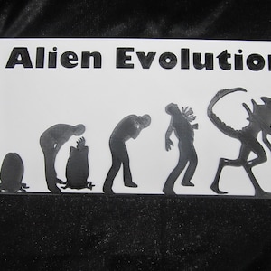 May include: A white rectangular sign with the words "Alien Evolution" in bold black letters. Below the text are black silhouettes depicting the stages of an alien's evolution, from egg to fully formed creature.