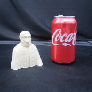 May include: A white, 3D printed bust of a primate wearing a robe. The bust is about the size of a standard can of soda.