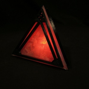 May include: A triangular, geometric object with a black frame and a red, illuminated interior. The object has a detailed pattern visible within the red light, and is set against a dark background.