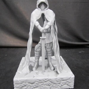 May include: A gray figurine of a robed figure holding a sword. The figure wears a crown and stands on a rectangular base with a textured surface. The figure's cloak drapes down the sides, and the sword is held upright.