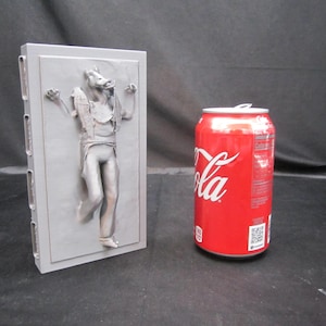 May include: A gray relief sculpture of a character in a frozen pose, mounted on a rectangular plaque. A red Coca-Cola can is next to the sculpture. The sculpture is a Star Wars themed item.