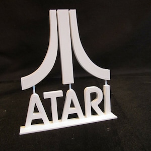 May include: White plastic 3D printed model of the Atari logo. The logo features the iconic Atari symbol and the word "ATARI" below it.