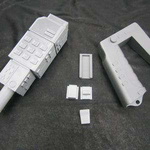 May include: A light gray, 3D-printed prop, possibly a futuristic device or a component of a larger machine. The image shows several detached parts, including a keypad, handle, and smaller rectangular pieces. The objects are arranged on a dark surface.