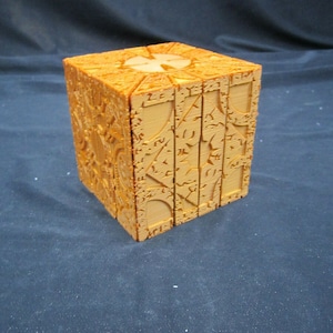 May include: A wooden cube puzzle with intricate carvings and symbols on each side. The cube is a light brown color and appears to be made of multiple layers.
