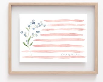 May include: Watercolor art print in a wooden frame. The artwork features a floral design with blue flowers and pink horizontal stripes, resembling a flag. The words "Land of the free" are written in script. The print is on white paper.