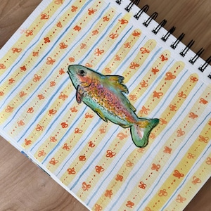 May include: A watercolor painting of a colorful fish on a spiral-bound sketchbook. The background features yellow and blue stripes with orange butterfly accents. The fish has a rainbow of colors, including green, red, and yellow.