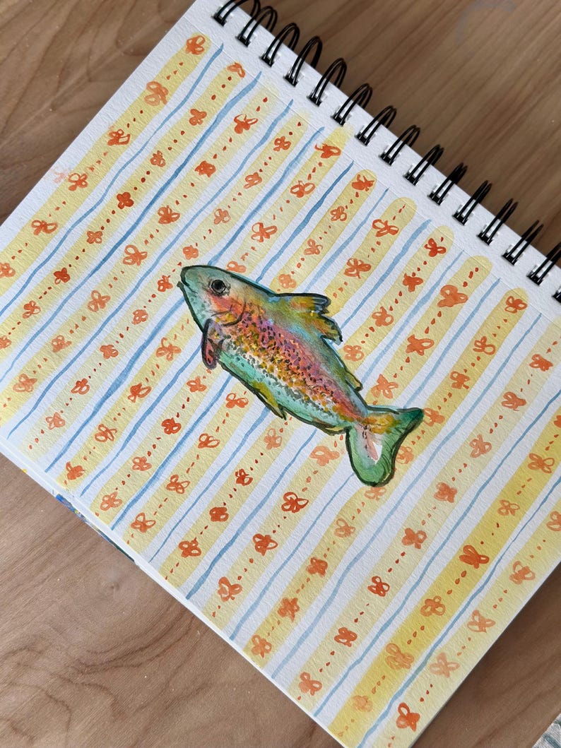 May include: A watercolor painting of a colorful fish on a spiral-bound sketchbook. The background features yellow and blue stripes with orange butterfly accents. The fish has a rainbow of colors, including green, red, and yellow.