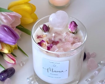May include: A lit candle in a clear glass jar with a white wax interior, decorated with rose quartz crystals, dried rose buds, and a heart-shaped crystal. The label reads "Mama". A small lit votive candle and flowers are in the background.