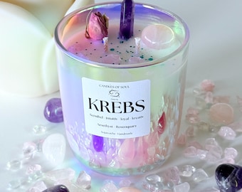 May include: A decorative candle in a glass jar with a holographic finish. The candle is adorned with crystals, a dried rose, and a label that reads "KREBS". Surrounding the candle are various crystals and stones in shades of pink, purple, and clear.