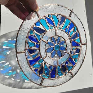 May include: Circular stained glass art piece with a floral design. The artwork features a combination of blue, turquoise, and clear glass pieces, framed by a copper-colored metal. The design includes a central flower and radiating patterns.
