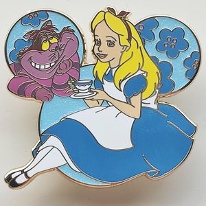 Op de afbeelding: Een Disney-pin met Alice in Wonderland. Alice, in haar blauwe jurk, houdt een kopje thee vast, met de Cheshire Cat naast haar. De pin heeft de vorm van Mickey Mouse-oren, met een lichtblauwe achtergrond en bloemaccenten.