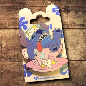 Op de afbeelding: Een Disney-pin met Stitch in een theekopje met Donald Duck. De pin heeft een lichtroze theekopje en schotel, met Stitch in een benarde pose. De pin zit op een witte achtergrond met blauwe bloemaccenten.