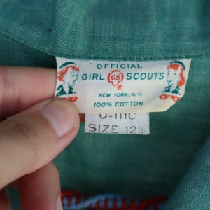 May include: Close-up of a green fabric with a label that reads "OFFICIAL GIRL SCOUTS NEW YORK, N.Y. 100% COTTON." The label also includes the size "12 1/2". The fabric is a solid green color.