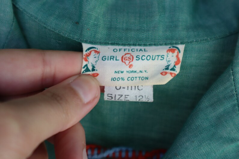 May include: Close-up of a green fabric with a label that reads "OFFICIAL GIRL SCOUTS NEW YORK, N.Y. 100% COTTON." The label also includes the size "12 1/2". The fabric is a solid green color.