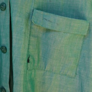 May include: Close-up of a teal green linen garment with a button-up front and a chest pocket. The fabric has a textured appearance. The buttons are a matching teal green color. The garment appears to be a shirt or jacket.