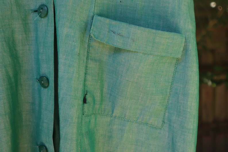 May include: Close-up of a teal green linen garment with a button-up front and a chest pocket. The fabric has a textured appearance. The buttons are a matching teal green color. The garment appears to be a shirt or jacket.