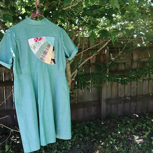 May include: A light green dress with short sleeves and a collared neckline hangs from a clothes hanger. The dress features a decorative patch on the back with floral and patterned fabric pieces. The dress is hanging outside near a wooden fence.