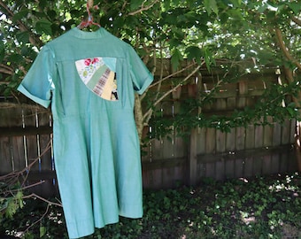 May include: A light green dress with short sleeves and a collared neckline hangs from a clothes hanger. The dress features a decorative patch on the back with floral and patterned fabric pieces. The dress is hanging outside near a wooden fence.