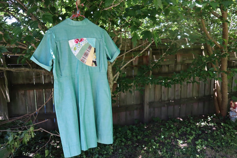 May include: A light green dress with short sleeves and a collared neckline hangs from a clothes hanger. The dress features a decorative patch on the back with floral and patterned fabric pieces. The dress is hanging outside near a wooden fence.