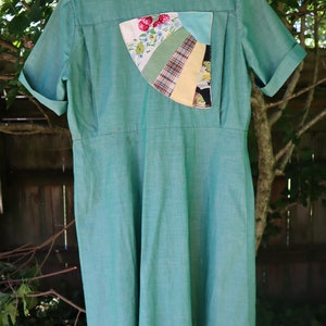 May include: A vintage-style, short-sleeved dress in a soft teal green. The dress features a unique patchwork detail on the back, incorporating floral, plaid, and solid fabric sections. The dress is hanging outdoors, showcasing its classic design and color.