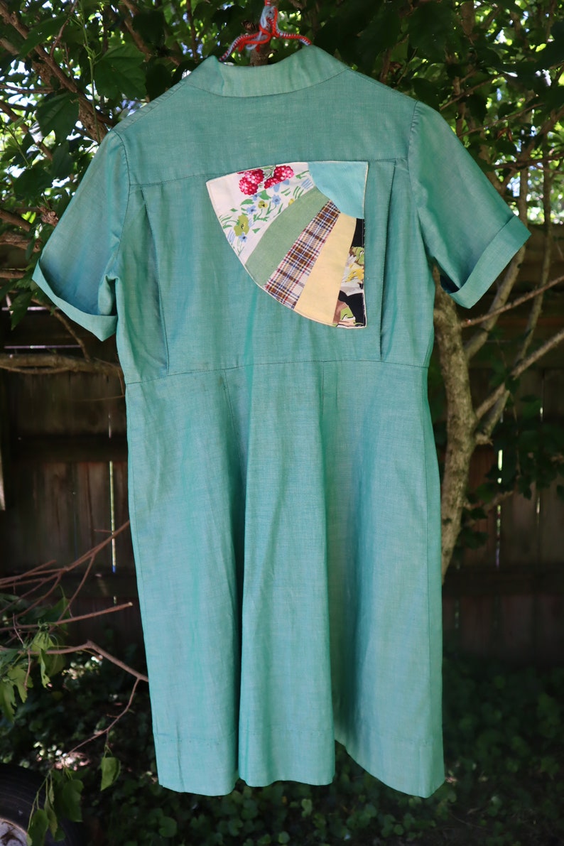 May include: A vintage-style, short-sleeved dress in a soft teal green. The dress features a unique patchwork detail on the back, incorporating floral, plaid, and solid fabric sections. The dress is hanging outdoors, showcasing its classic design and color.