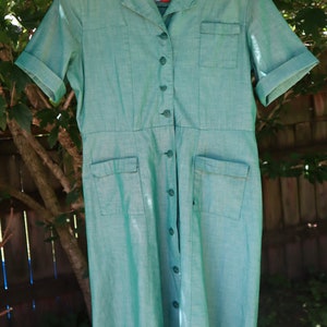May include: A vintage turquoise dress with short sleeves, a collared neckline, and a button-down front. The dress features two chest pockets and two hip pockets. The dress is hanging on a red and white hanger.