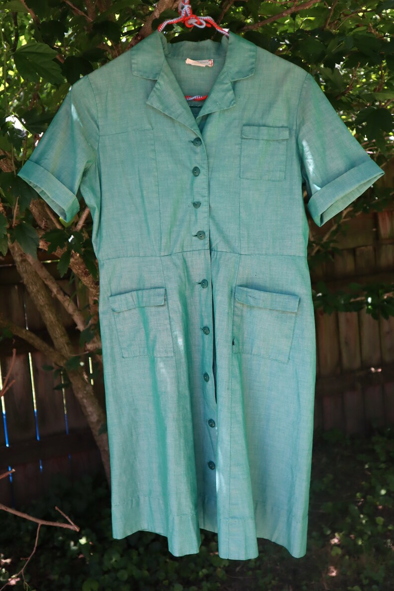 May include: A vintage turquoise dress with short sleeves, a collared neckline, and a button-down front. The dress features two chest pockets and two hip pockets. The dress is hanging on a red and white hanger.