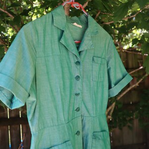 May include: A vintage green dress with short sleeves, a collared neckline, and button-down front. The dress has two front pockets and is hanging on a clothes hanger against a backdrop of green foliage and a wooden fence.