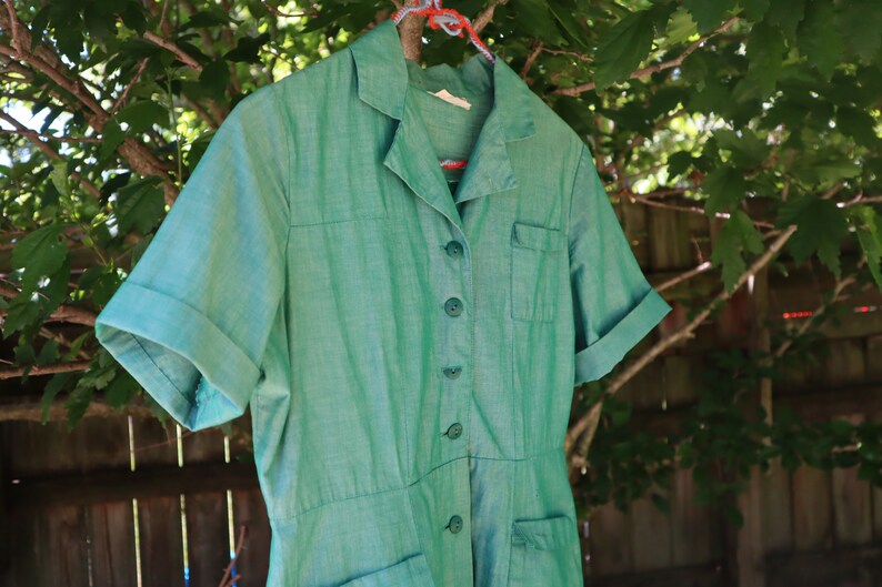 May include: A vintage green dress with short sleeves, a collared neckline, and button-down front. The dress has two front pockets and is hanging on a clothes hanger against a backdrop of green foliage and a wooden fence.