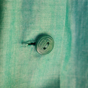 May include: Close-up of a teal fabric with a button and buttonhole. The button is round, translucent, and has a decorative design. The fabric has a subtle texture and is a solid teal color.