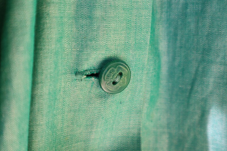 May include: Close-up of a teal fabric with a button and buttonhole. The button is round, translucent, and has a decorative design. The fabric has a subtle texture and is a solid teal color.