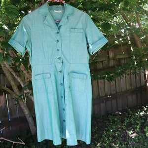 May include: A vintage turquoise dress with short sleeves, a collared neckline, and button-down front. The dress features two front pockets and a fitted waist. The dress is hanging on a pink hanger outdoors, with a wooden fence and greenery in the background.
