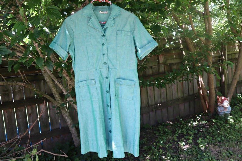 May include: A vintage turquoise dress with short sleeves, a collared neckline, and button-down front. The dress features two front pockets and a fitted waist. The dress is hanging on a pink hanger outdoors, with a wooden fence and greenery in the background.