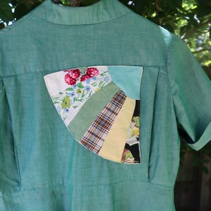 May include: A teal-green collared shirt with short, cuffed sleeves. The back features a unique patchwork pocket with floral, plaid, and solid fabric segments. The shirt has a vintage aesthetic.