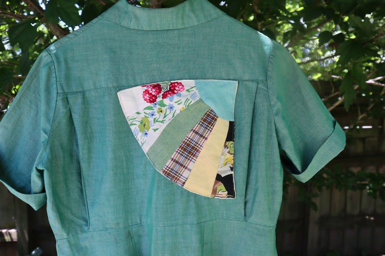 May include: A teal-green collared shirt with short, cuffed sleeves. The back features a unique patchwork pocket with floral, plaid, and solid fabric segments. The shirt has a vintage aesthetic.