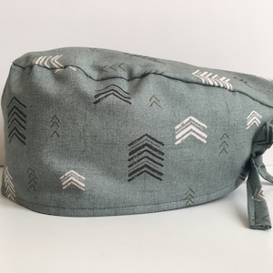 May include: A teal scrub cap with a repeating pattern of black, white, and light brown arrows. The cap has a tie closure and is made of a soft, textured fabric. The arrows are arranged in various directions.