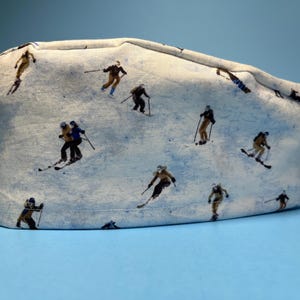 May include: A light blue surgical cap featuring a repeating pattern of skiers on a snowy mountain slope. The skiers are depicted in various poses, with brown and yellow outfits, skiing down the mountain. The cap has a tie closure.