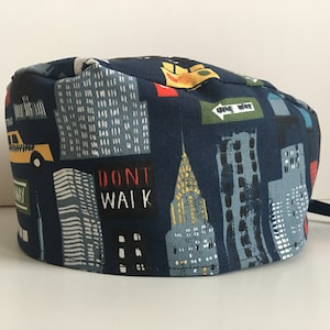 May include: A navy blue scrub cap featuring a cityscape design. The cap is patterned with illustrations of skyscrapers, yellow taxis, and street signs. The words "DON'T WALK" and "ONE WAY" are visible on the cap.