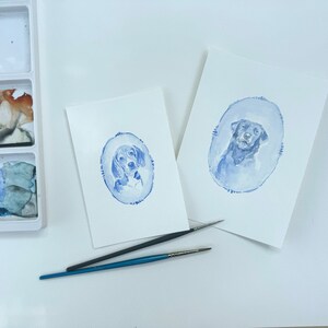 May include: Two watercolor paintings of dogs on white paper. The paintings are in blue tones and feature a dog in an oval frame. The paintings are on a white surface with two paintbrushes.