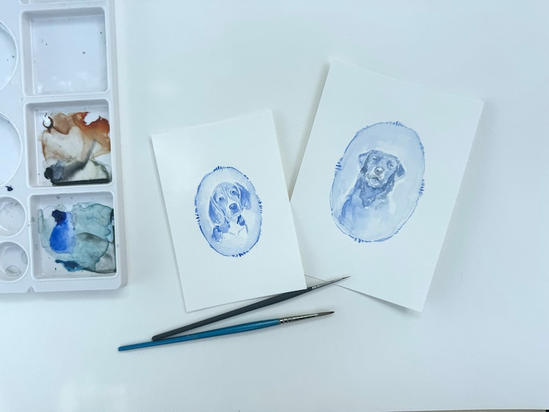 May include: Two watercolor paintings of dogs on white paper. The paintings are in blue tones and feature a dog in an oval frame. The paintings are on a white surface with two paintbrushes.