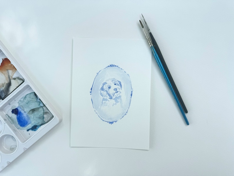 May include: A watercolor painting of a dog's head in a blue oval frame. The dog is white with a light blue background.