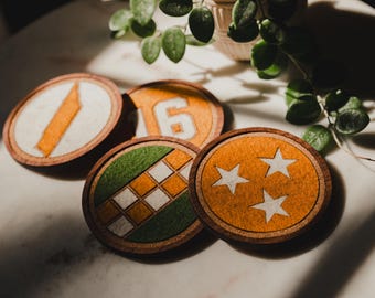 Op de afbeelding: Een verzameling ronde houten onderzetters met verschillende ontwerpen. Eén onderzetter heeft een oranje streep, een andere het nummer 6, een derde een groen-wit geblokt patroon en de laatste heeft drie witte sterren op een oranje achtergrond.