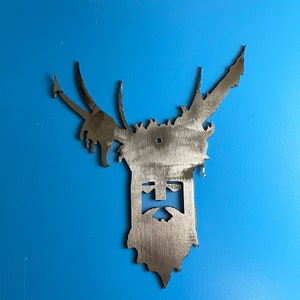 Puede incluir: Un recorte de metal de una cara con cuernos. La cara tiene barba y un diseño en forma de cruz en el centro.