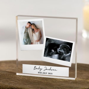 Op de afbeelding: Een helder acryl fotoblok met twee afgedrukte foto's: een stel en een echo. Het blok bevat de tekst "Baby Jackson DUE JULY 2025". Het staat op een houten oppervlak.