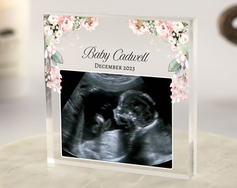 May include: A clear acrylic block with a floral design border and a black and white ultrasound image inside. The text "Baby Cadwell December 2023" is printed on the block.