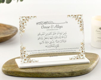 May include: A white acrylic plaque with a floral design and the text "Omar & Aliya 30th March 2024" and a Quran verse in Arabic script. The plaque is on a white stand and a wooden surface.