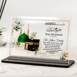 Könnte beinhalten: Eine klare Acrylplatte mit goldener Glitzerbordüre und einer Aquarell-Illustration der Kaaba und der Masjid al-Nabawi. Der Text "Hajj Mubarak Juni 2024" und "The Khan Family" ist auf der Platte gedruckt. Die Platte steht auf einem schwarzen Ständer.